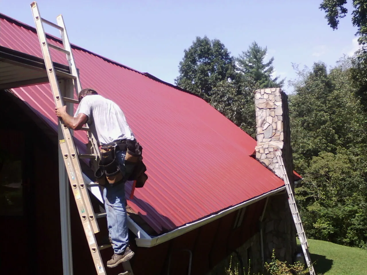 Roofer on a ladder beside a red metal roof and stone chimney — Soffit & Fascia Repair by Roofing Brothers in Miamitown